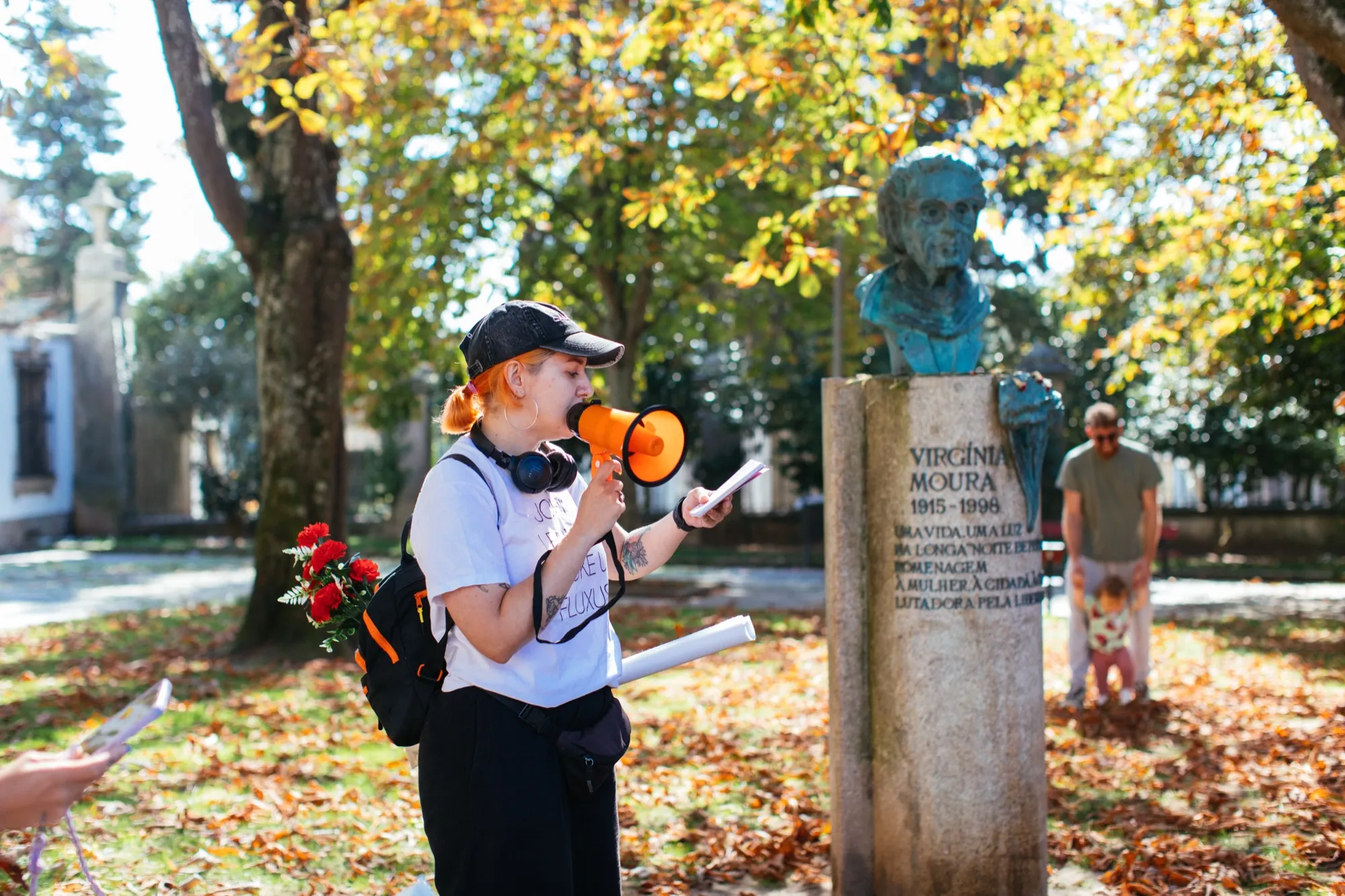 A guia, com megafone, junto ao busto de Virgínia Moura, entre rosas e folhas de outono.