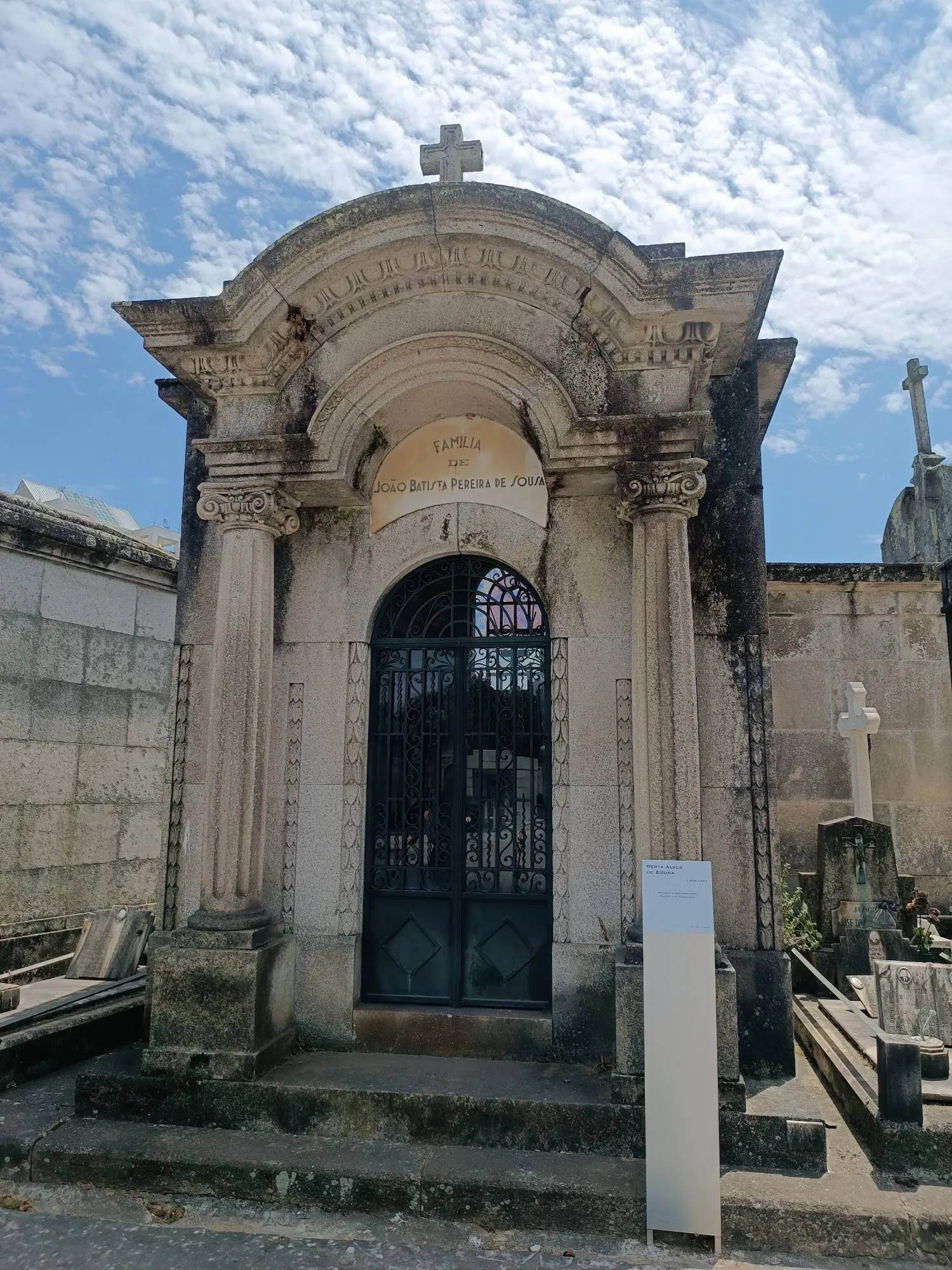 Um jazigo-capela neoclássico, com colunas e porta de ferro, sob um céu de nuvens.